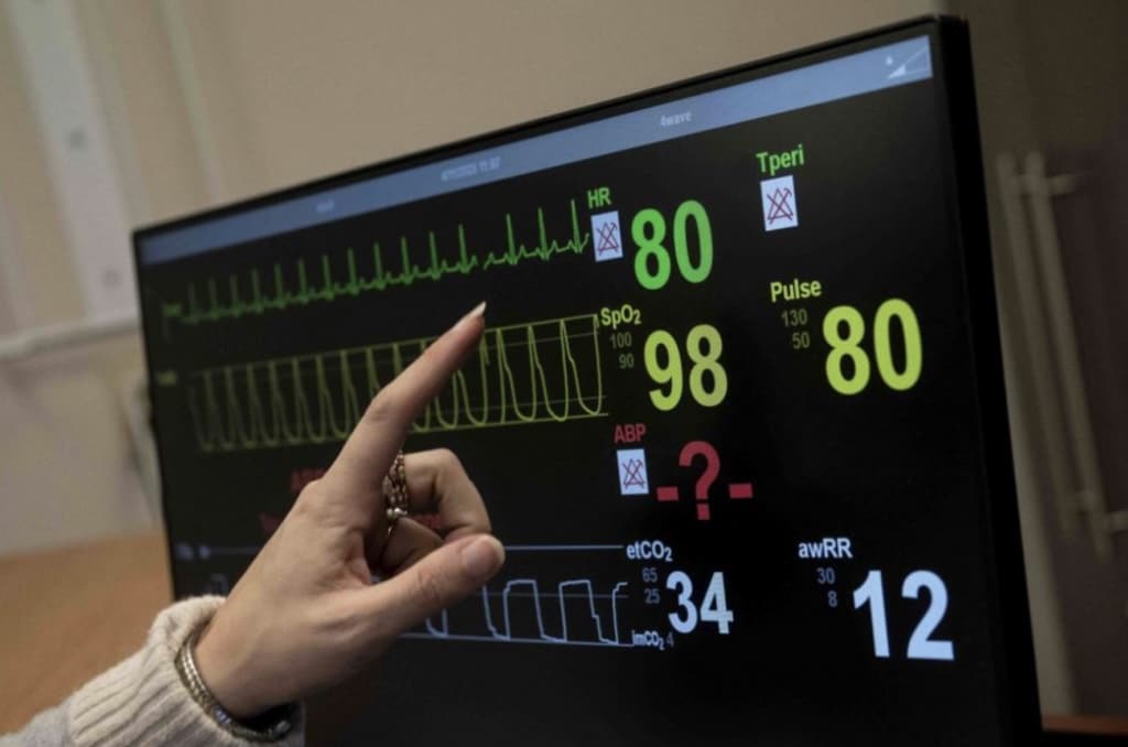 A hand points at a medical monitor during a nursing simulation, displaying vital signs—heart rate (80), oxygen saturation (98), pulse (80)—with some measurements marked with red "X" for missing or abnormal values at Bryant & Stratton College.